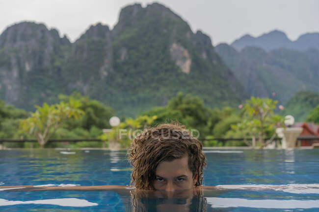 Attraente giovane uomo che guarda la macchina fotografica e nuota in piscina sullo sfondo di verdi colline. — Foto stock