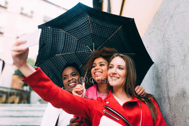Grupo de tres hermosas mujeres jóvenes multirraciales divirtiéndose caminando sobre la lluvia - foto de stock