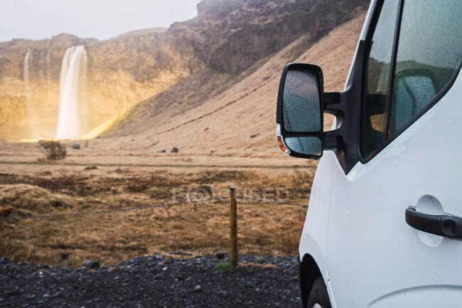 Kornfragment eines auf dem Boden geparkten weißen Fahrzeugs mit Blick auf Berge und Wasserfall im Hintergrund, Island. — Stockfoto