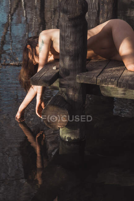 Senhora nua com tatuagens no cais de madeira com a mão no rio na natureza — Fotografia de Stock