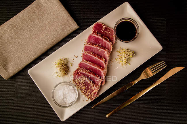 Raw sliced tuna steak on platter with sauce on black background — Stock Photo