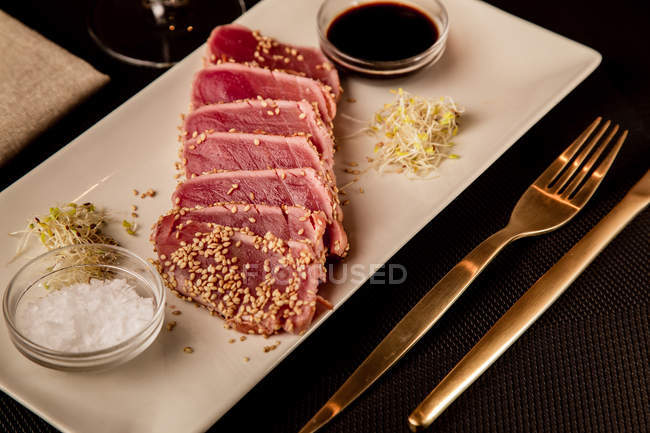 Сырой нарезанный стейк из тунца на блюдечке с соусом на черном фоне — стоковое фото