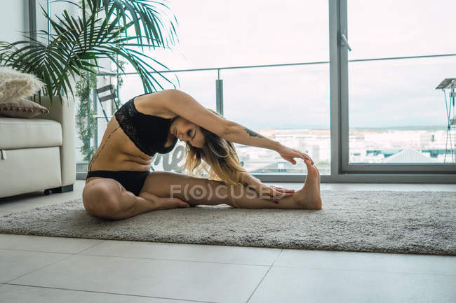 Confident tattooed woman in black lingerie sitting on carpet against window and practicing yoga asana — Stock Photo