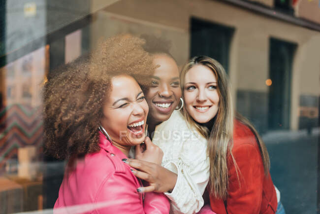 Grupo de tres hermosas mujeres jóvenes multirraciales sentadas en una cafetería mirando a la ventana abrazándose - foto de stock