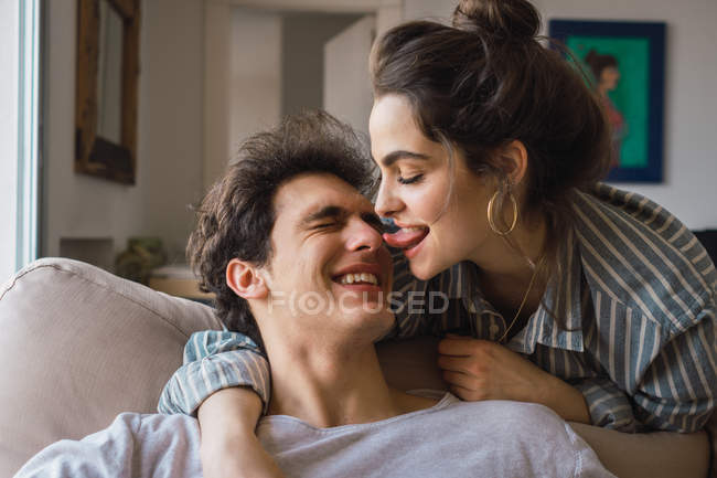 Mulher alegre lambendo nariz de homem sorridente no sofá em casa — Fotografia de Stock