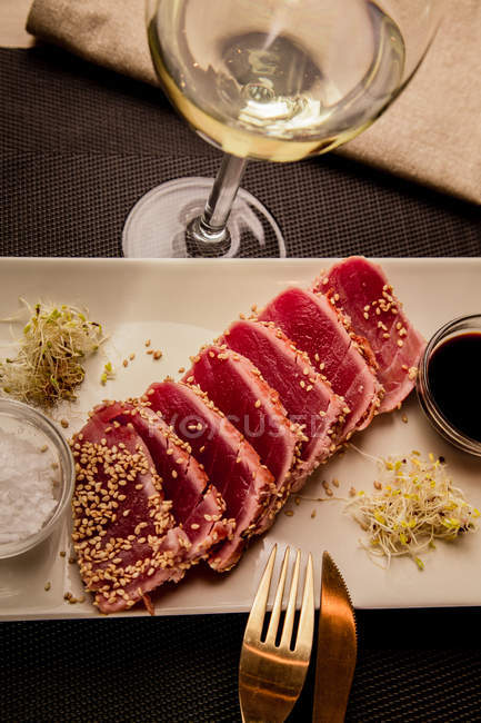 Сырой нарезанный стейк из тунца на блюдечке с соусом на черном фоне с бокалом белого вина — стоковое фото
