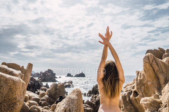 Back view of naked woman with hands up standing and stretching at coastal rocks at the ocean. — Stock Photo