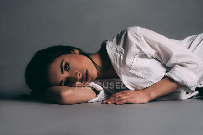 Mulher morena atraente na camisa deitada com a cabeça na mão olhando para a câmera no fundo cinza — Fotografia de Stock