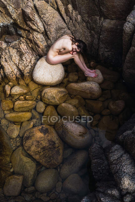 Dall'alto donna nuda seduta con le mani dietro la schiena su pietre. — Foto stock