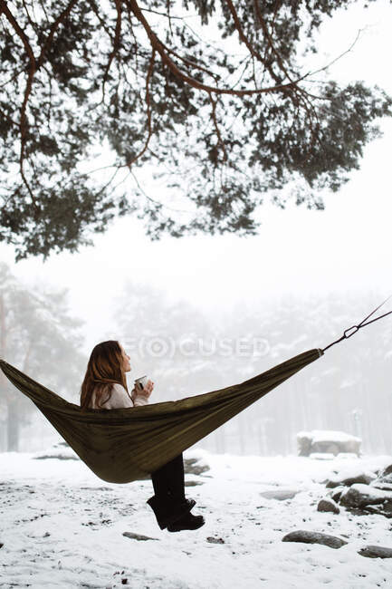 Вид збоку на досить мрійливу молоду жінку, яка сидить в гамаку і має гарячий напій в зимовій природі . — стокове фото