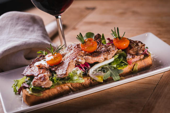 Sandwich with fried meat and vegetables and glass of red wine on wooden table — Stock Photo
