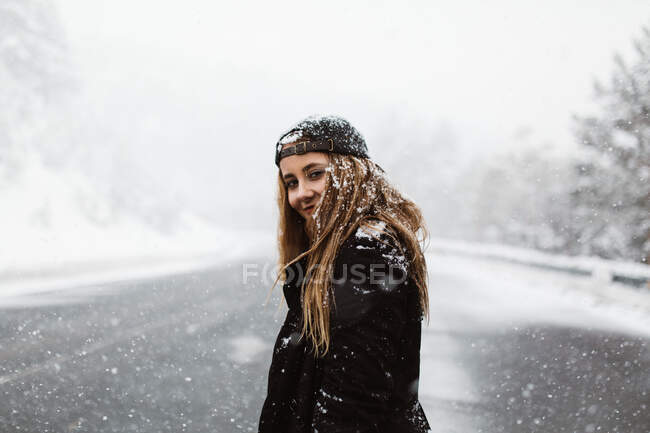 Vue latérale de jolie femme debout et regardant la caméra sur la route dans les chutes de neige. — Photo de stock