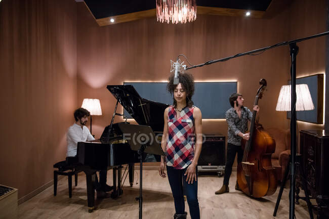 Lovely woman in casual outfit singing nice song white rehearsing with band in recording studio. — Stock Photo