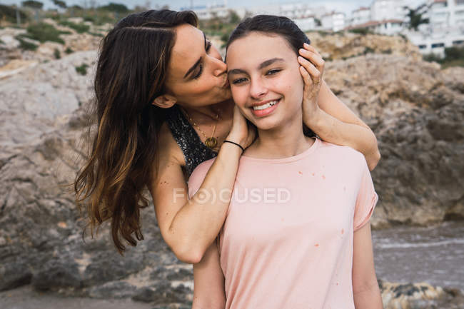 Жінка обіймає і цілує усміхнену дочку на морському фоні влітку — стокове фото