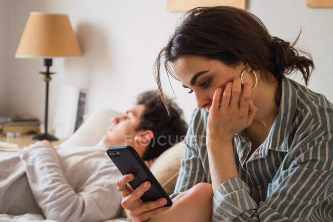 Giovane donna triste seduta e utilizzando smartphone mentre il fidanzato dorme sul letto — Foto stock