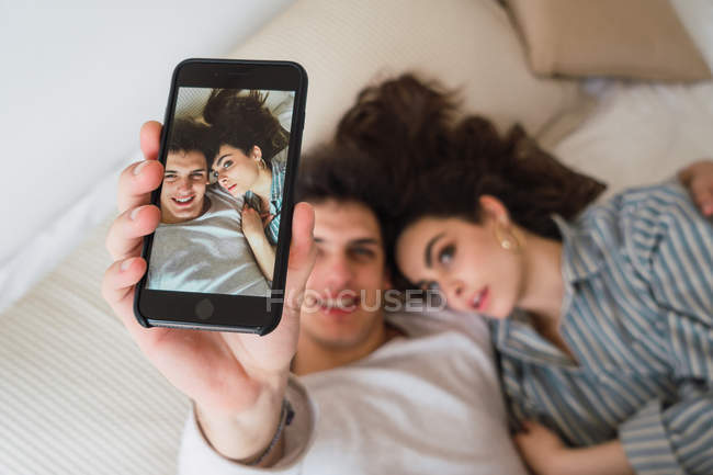 Alegre joven pareja tomando selfie con smartphone en la cama - foto de stock