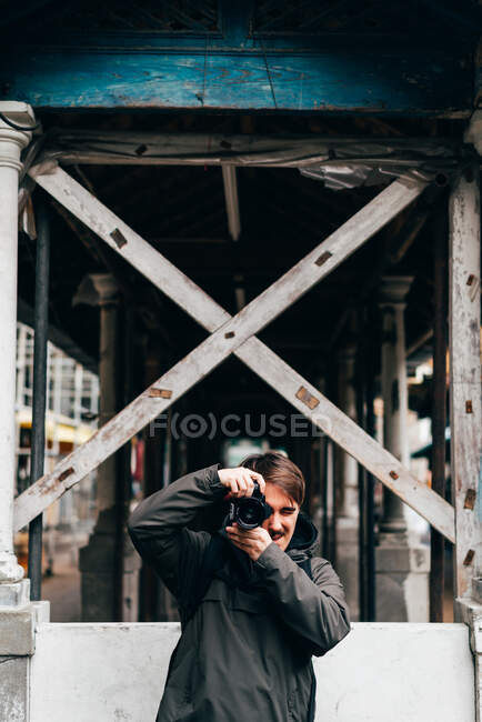 Jovem fotógrafo homem tomando tiros sob construção de madeira na rua da cidade. — Fotografia de Stock