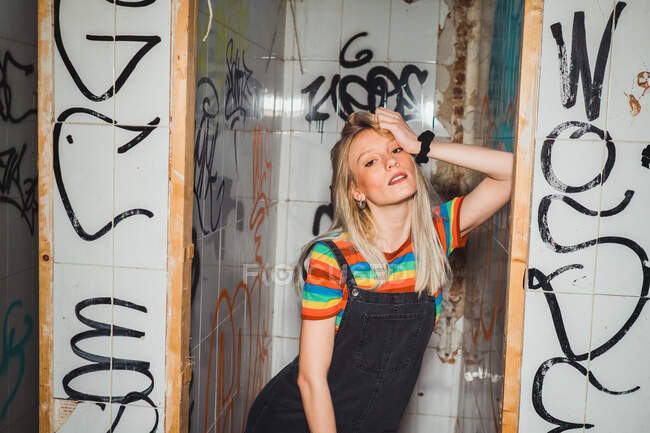 Young blond model in denim and colorful t-shirt standing provocatively in abandoned restroom with graffiti on wall. — Stock Photo