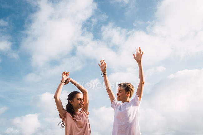Смеющийся мальчик и девочка стоят с поднятыми вверх руками перед небом — стоковое фото