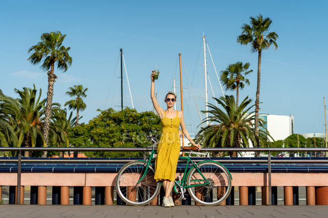 Frau in sommergelbem Kleid lehnt auf Fahrrad und hält Getränk hoch — Stockfoto