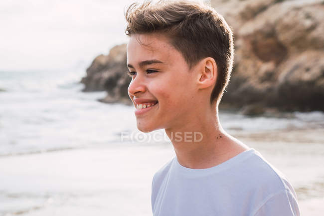Smiling cute boy in white t-shirt standing on seashore in summer — Stock Photo