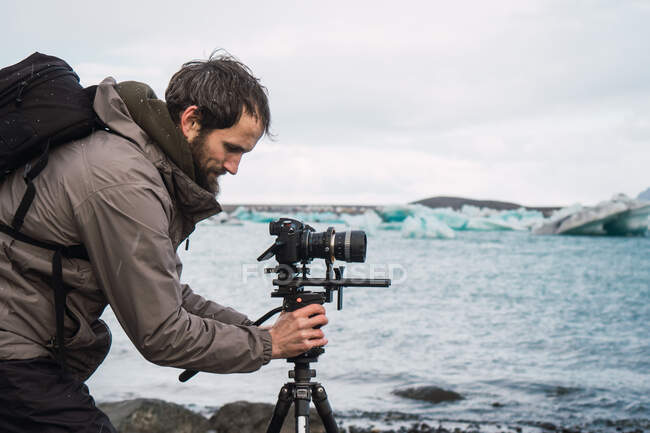 Vista laterale dell'uomo in outwear con zaino impostazione fotocamera fotografica su treppiede per scattare foto di bel paesaggio marino freddo. — Foto stock