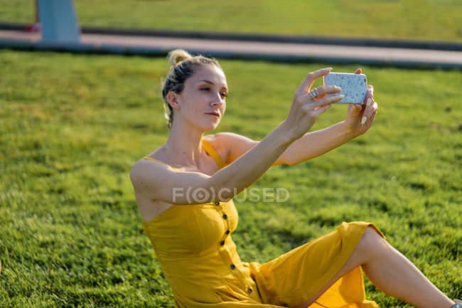 Mujer sentada en la hierba en la ciudad con smartphone y tomando selfie - foto de stock