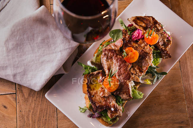 Sandwich with fried meat and vegetables and glass of red wine on wooden table — Stock Photo