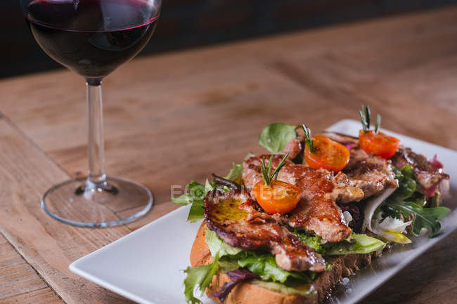 Sanduíche com carne frita e legumes e copo de vinho tinto na mesa de madeira — Fotografia de Stock