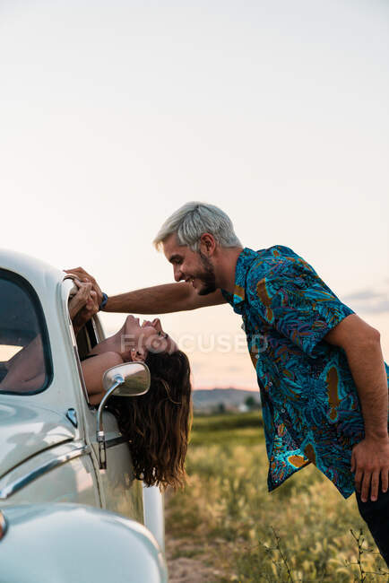 Side view of woman sitting inside of car leaning head out and looking at man standing above and smiling happily in summertime — Stock Photo