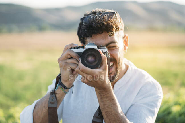 Поганий молодий хлопець у сонцезахисних окулярах радісно посміхається і тримає фотокамеру ретро, стоячи на розмитому фоні дивовижної сільської місцевості. — стокове фото