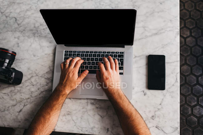 Crop shot from above of man using laptop sitting at marble table with phone near — Stock Photo