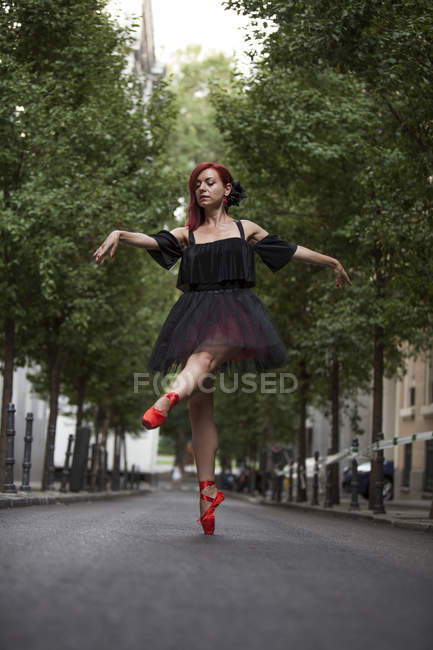 Рыжая балерина с черной пачкой и красными балетными кончиками танцует на улице с деревьями на заднем плане . — стоковое фото