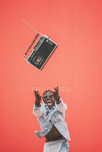 Juguetón afroamericano hombre lanzando dispositivo de radio vintage sobre fondo rojo - foto de stock