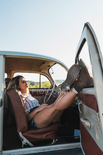 Schlafende Frau lehnt Beine an geöffnete Tür des Retro-Autos und schläft auf Sitz im Sonnenlicht — Stockfoto