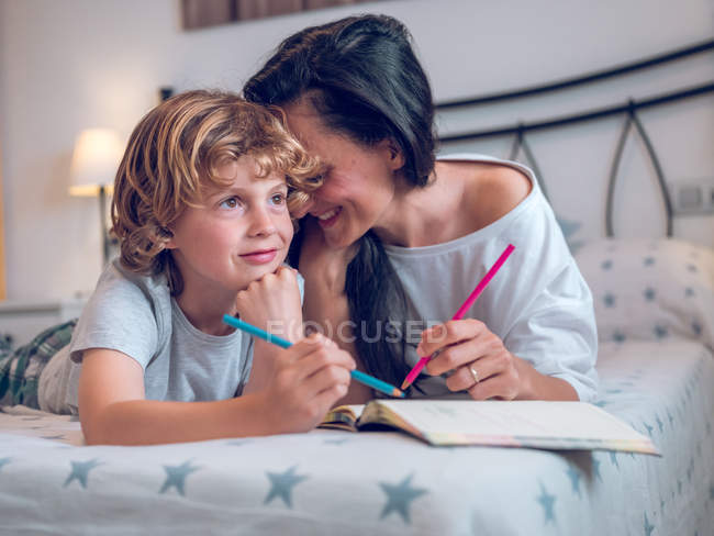 Beautiful woman and cute child lying on comfortable bed and coloring pictures in notebook together — стокове фото