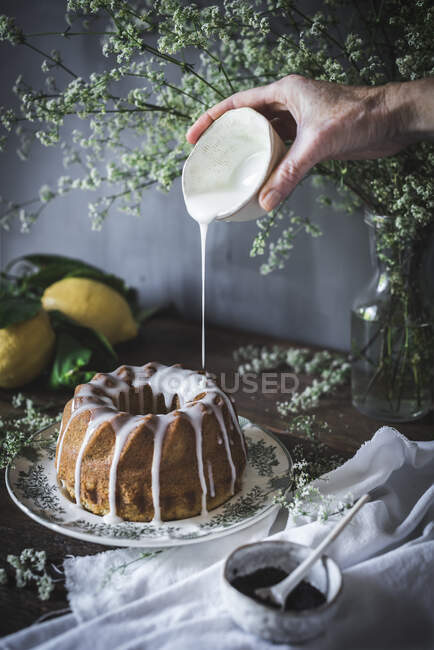 Crop view of hand holding cup of sweet dressing and pouring on delicious pie placed on dish near cup of poppy seed and flowers in vase — Stock Photo