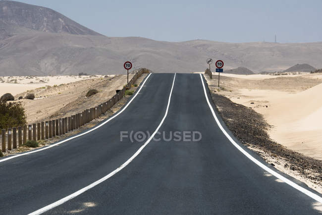 Estrada reta limpa com sinais em planície arenosa seca com colinas e montanhas, Ilhas Canárias — Fotografia de Stock