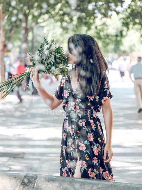 Attractive brunette woman in dark dress standing on street and smelling fresh beautiful flowers on sunny street — Stock Photo