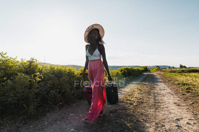 Donna afro-americana alla moda in cappello che porta valigia e cammina su strada rurale in estate — Foto stock