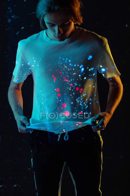 Андрогінна чоловіча модель дивиться на яскраві плями червоного і синього світла на футболці, стоячи на чорному тлі. — стокове фото