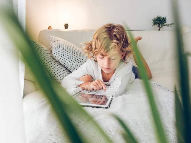 Мальчик пользуется цифровым планшетом, лежа на диване дома — стоковое фото