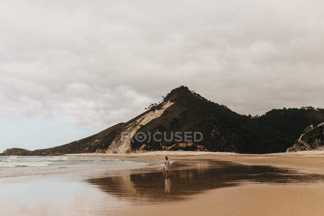 Persona caminando en la orilla arenosa húmeda cerca del agua de mar en el fondo de la montaña y el cielo nublado - foto de stock
