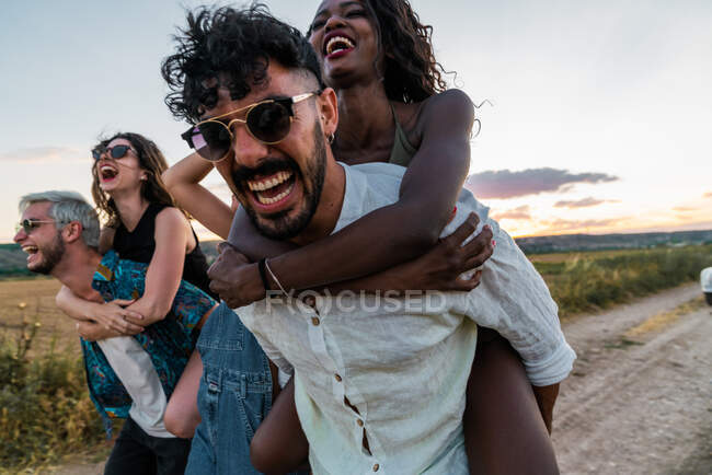 Two men giving piggyback ride to women and running along countryside road while spending time in nature together — Stock Photo