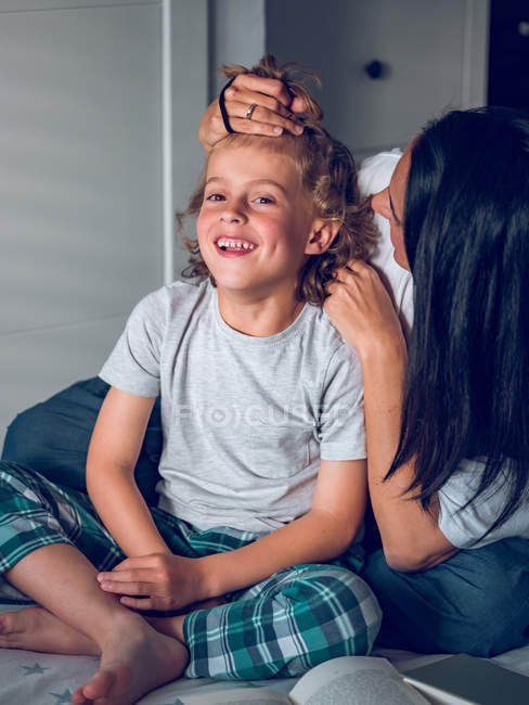 Mulher adulta sentada na cama atrás de menino alegre e montando seu cabelo na cauda — Fotografia de Stock