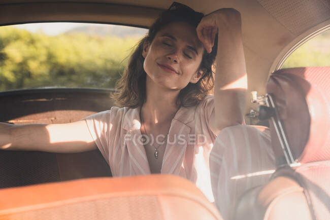 Hermosa mujer confiada en rosa y gafas de sol sentado en el asiento trasero en coche retro mirando hacia otro lado a la luz del sol - foto de stock