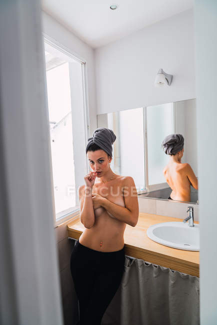 Junge Oben-ohne-Frau in schwarzen Strumpfhosen und Handtuch auf dem Kopf, die im Badezimmer am Fenster steht und die Brust mit Armen bedeckt — Stockfoto