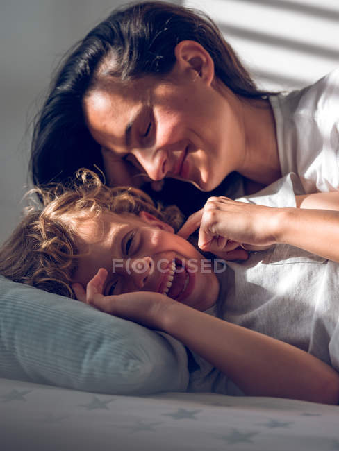 Mulher bonita deitada na cama atrás do menino doce e tocando sua bochecha com cuidado — Fotografia de Stock