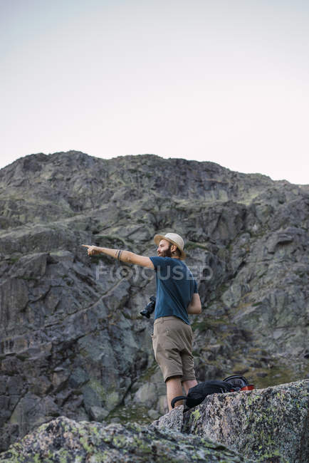 Young man in hat standing on rock in mountains and holding photo camera — Stock Photo