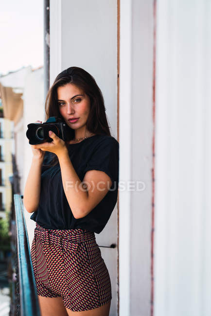 Молода вдумлива жінка стоїть на балконі з фотоапаратом — стокове фото